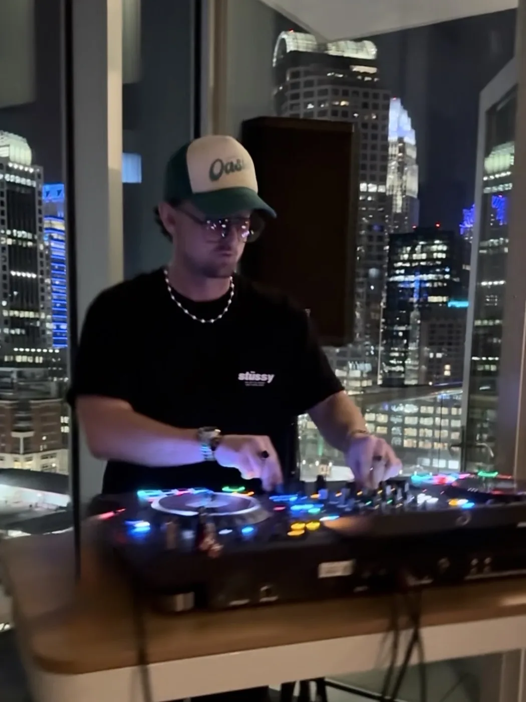 AUSTAL DJing in a high-rise apartment at night, city skyline through the window